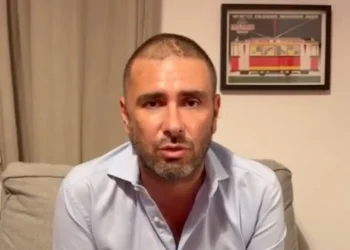 Alessandro Di Battista seduto su un divano grigio, camicia azzurra, con una cicatrice visibile sul capo rasato