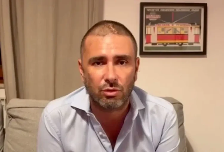 Alessandro Di Battista seduto su un divano grigio, camicia azzurra, con una cicatrice visibile sul capo rasato