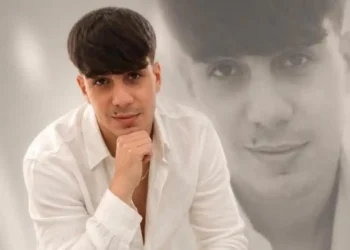 Alessio Giuga, 22 anni, di Rosolini, in camicia bianca con la mano sotto il mento, davanti a un suo ritratto in bianco e nero