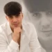 Alessio Giuga, 22 anni, di Rosolini, in camicia bianca con la mano sotto il mento, davanti a un suo ritratto in bianco e nero