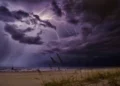 Temporale con fulmini sopra il mare, nubi scure, pioggia al largo e una barca a vela vicino alla riva di una spiaggia