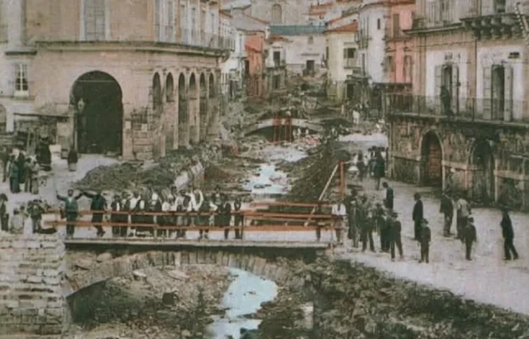 Modica dopo l'alluvione del 1902: alveo pieno di detriti tra i palazzi, gente su una passerella di legno e sulle strade