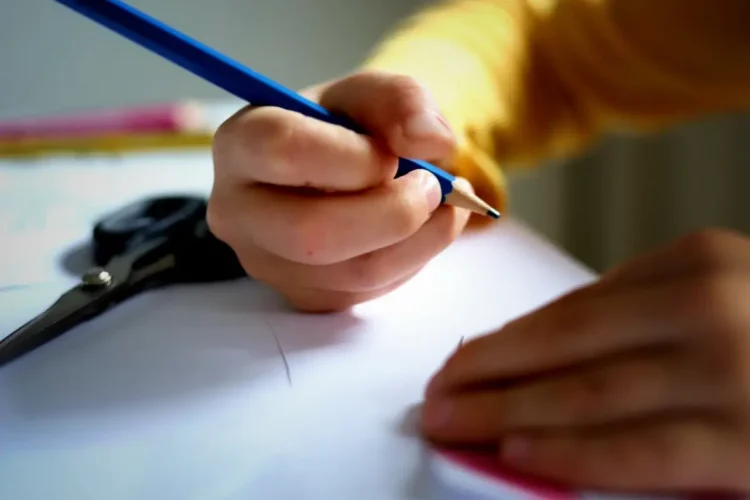 Bambino scrive con una matita blu su un foglio bianco, alle porte del nuovo anno scolastico