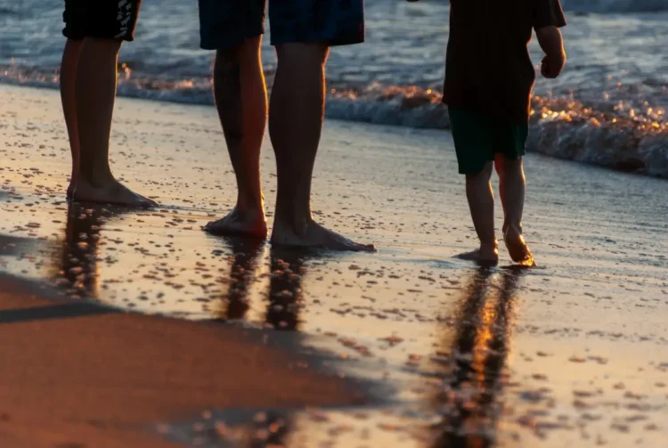 Due adulti e un bambino camminano a piedi nudi sulla battigia al tramonto