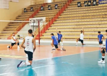 Giocatori dell'Avimec Modica si allungano a bordo campo durante il riscaldamento in palestra, spalti sullo sfondo