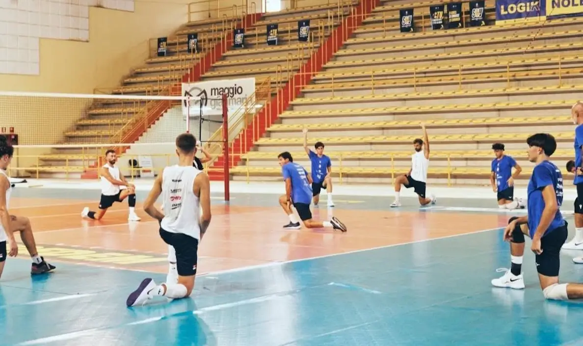Giocatori dell'Avimec Modica si allungano a bordo campo durante il riscaldamento in palestra, spalti sullo sfondo