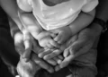 Mani di due genitori che sorreggono le mani di un bambino con la maglietta bianca, foto in bianco e nero sul tema famiglie