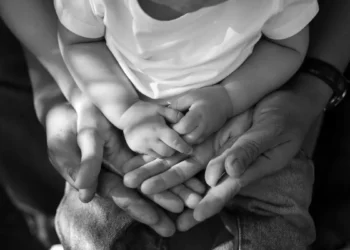 Mani di due genitori che sorreggono le mani di un bambino con la maglietta bianca, foto in bianco e nero sul tema famiglie