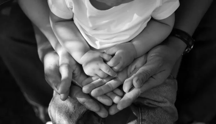 Mani di due genitori che sorreggono le mani di un bambino con la maglietta bianca, foto in bianco e nero sul tema famiglie
