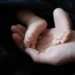 Piedini di un neonato appoggiati nel palmo della mano di un adulto, su un tessuto scuro