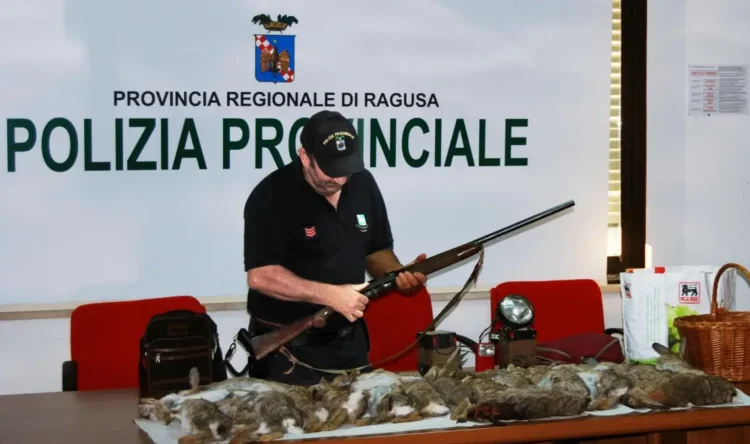 Agente della Polizia Provinciale di Ragusa esamina un fucile; sul tavolo selvaggina abbattuta e un faro portatile