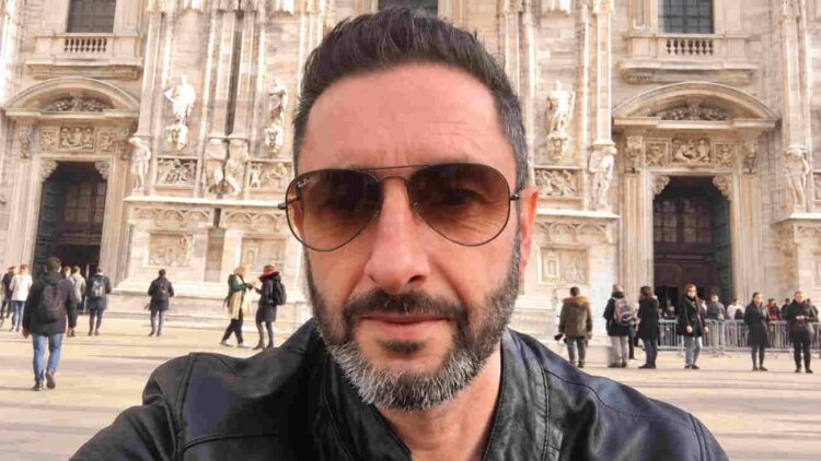 Uomo con barba brizzolata e occhiali da sole, ritratto all'aperto davanti a un edificio monumentale.