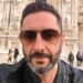 Uomo con barba brizzolata e occhiali da sole, ritratto all'aperto davanti a un edificio monumentale.
