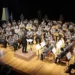 Orchestra Fiati in divisa grigia diretta da un maestro sul palco di un teatro, legata all'intesa tra Conservatorio Bellini e Ispica
