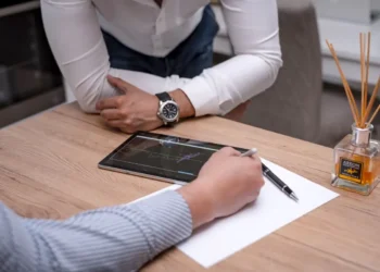 Una persona scrive su un foglio con la penna accanto a un tablet con un grafico, mentre un'altra si appoggia al tavolo