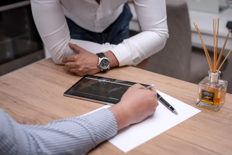 Una persona scrive su un foglio con la penna accanto a un tablet con un grafico, mentre un'altra si appoggia al tavolo