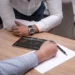 Una persona scrive su un foglio con la penna accanto a un tablet con un grafico, mentre un'altra si appoggia al tavolo
