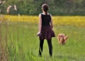 Una donna vista di spalle con il guinzaglio in mano cammina in un prato fiorito, poco dietro al suo cane dal pelo fulvo