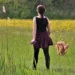 Una donna vista di spalle con il guinzaglio in mano cammina in un prato fiorito, poco dietro al suo cane dal pelo fulvo