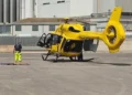 Elicottero giallo dell'elisoccorso fermo su un'elisuperficie asfaltata, con un soccorritore in piedi accanto a una barella a terra