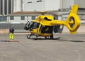 Elicottero giallo dell'elisoccorso fermo su un'elisuperficie asfaltata, con un soccorritore in piedi accanto a una barella a terra