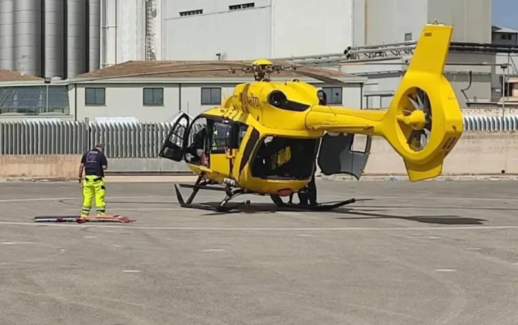 Elicottero giallo dell'elisoccorso fermo su un'elisuperficie asfaltata, con un soccorritore in piedi accanto a una barella a terra
