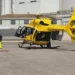 Elicottero giallo dell'elisoccorso fermo su un'elisuperficie asfaltata, con un soccorritore in piedi accanto a una barella a terra
