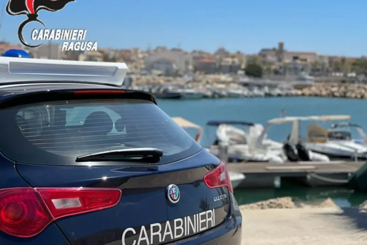 Auto dei Carabinieri di Ragusa ferma su una banchina, con un porticciolo e barche ormeggiate sullo sfondo