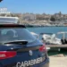 Auto dei Carabinieri di Ragusa ferma su una banchina, con un porticciolo e barche ormeggiate sullo sfondo