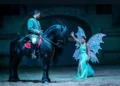 Spettacolo equestre: cavaliere in costume verde su un cavallo nero e una donna con ali da fata illuminate nell'arena
