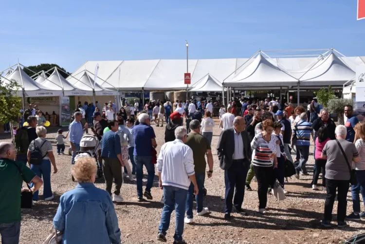 Visitatori tra i padiglioni bianchi della fiera agroalimentare di Ragusa in una giornata di sole, con balle di fieno sullo sfondo