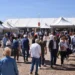 Visitatori tra i padiglioni bianchi della fiera agroalimentare di Ragusa in una giornata di sole, con balle di fieno sullo sfondo