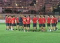 Giocatori del Frigintini in maglia rossa e pantaloncini blu in cerchio attorno all'allenatore su un campo sintetico, in notturna