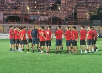 Giocatori del Frigintini in maglia rossa e pantaloncini blu in cerchio attorno all'allenatore su un campo sintetico, in notturna