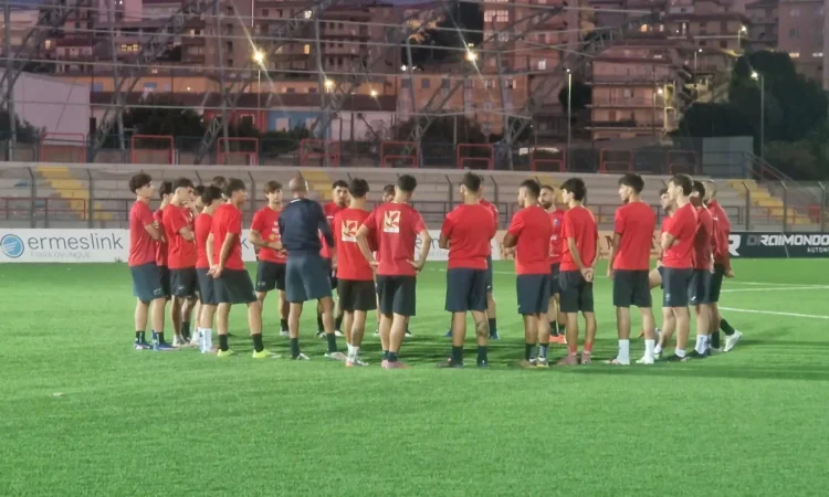 Giocatori del Frigintini in maglia rossa e pantaloncini blu in cerchio attorno all'allenatore su un campo sintetico, in notturna
