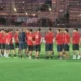 Giocatori del Frigintini in maglia rossa e pantaloncini blu in cerchio attorno all'allenatore su un campo sintetico, in notturna