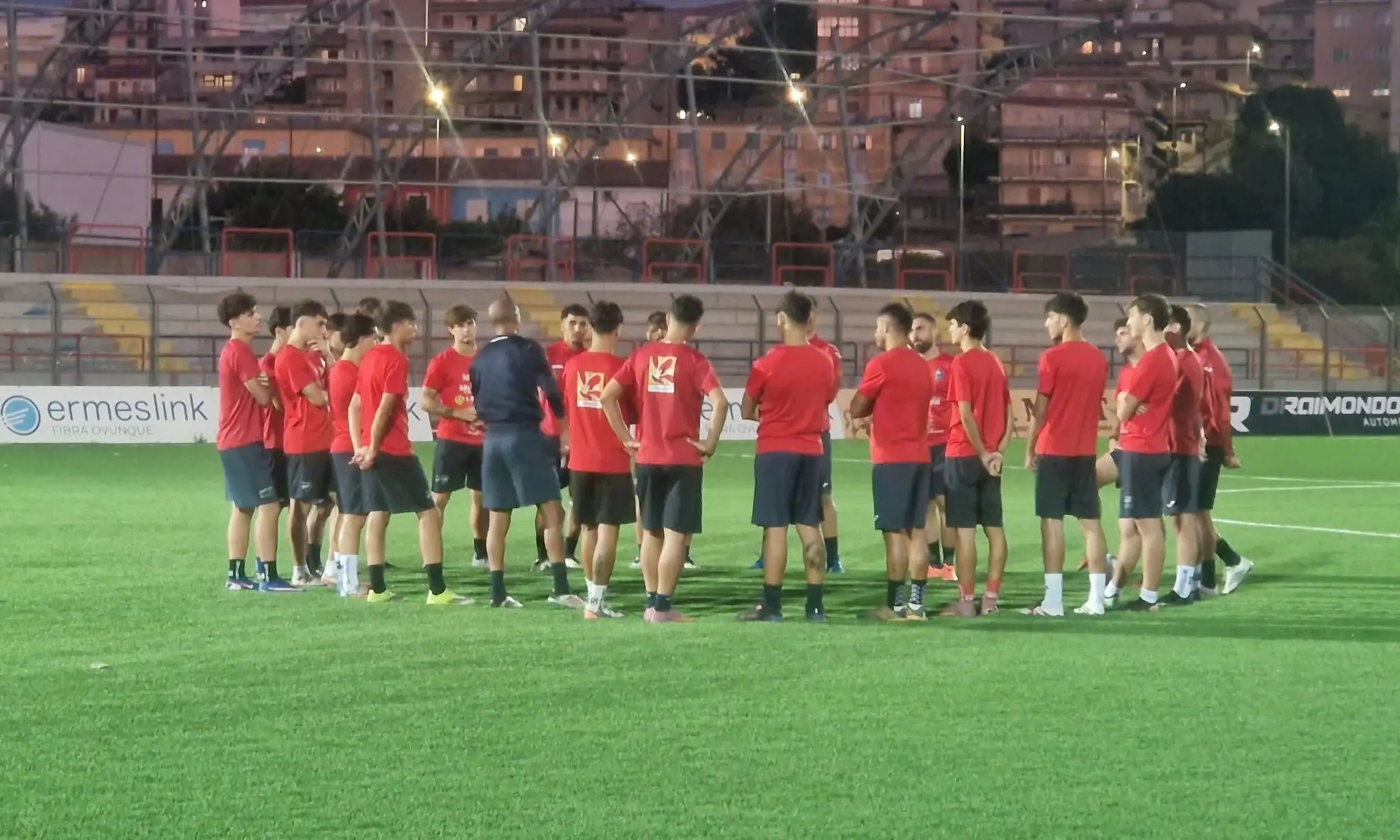 Giocatori del Frigintini in maglia rossa e pantaloncini blu in cerchio attorno all'allenatore su un campo sintetico, in notturna