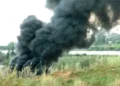 Fumarola di fumo nero denso che si leva da un campo tra erba alta, con strutture bianche e alberi sullo sfondo