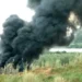 Fumarola di fumo nero denso che si leva da un campo tra erba alta, con strutture bianche e alberi sullo sfondo