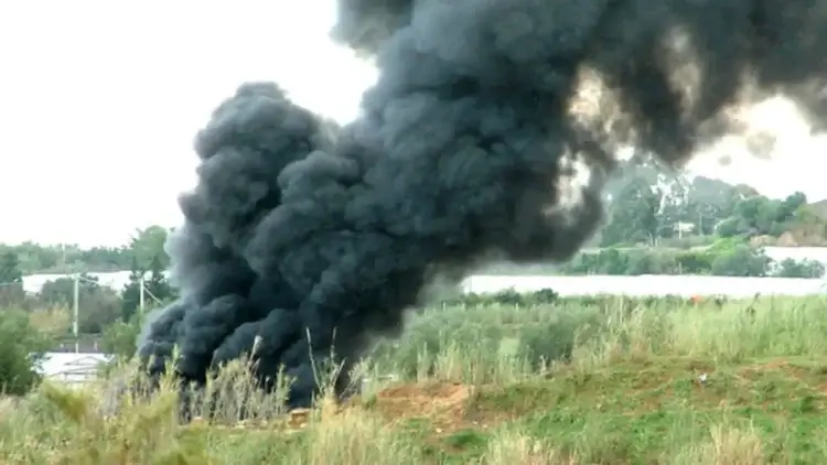 Fumarola di fumo nero denso che si leva da un campo tra erba alta, con strutture bianche e alberi sullo sfondo
