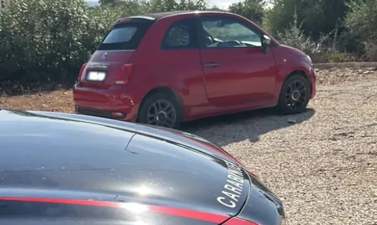 Fiat 500 rossa ferma su uno sterrato, accanto al cofano di un'auto dei Carabinieri