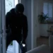 Uomo incappucciato con torcia entra da una porta socchiusa in un'abitazione buia, di notte
