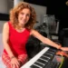 Grazia Colombo sorride al pianoforte, capelli ricci rossi, con un fiore rosso arancio sul leggio dello strumento
