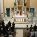 Messa vista dall'alto: celebranti all'altare, fedeli in piedi e, sulla destra, la statua dell'Immacolata