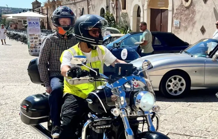 Due persone in sella a una moto con caschi integrali davanti al Castello di Donnafugata, a Ragusa, tra auto d'epoca