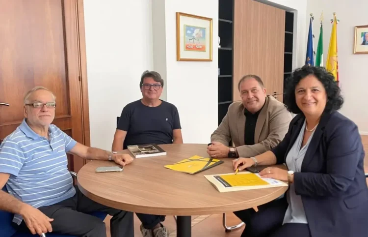 Quattro persone a un tavolo rotondo con opuscoli gialli del progetto In viaggio con i Cabrera, bandiere sullo sfondo