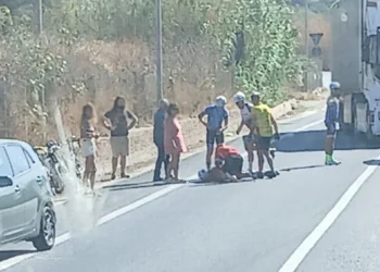 Un ciclista a terra soccorso da altri ciclisti e passanti lungo una strada provinciale, con auto e mezzo pesante in transito.