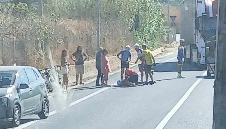 Un ciclista a terra soccorso da altri ciclisti e passanti lungo una strada provinciale, con auto e mezzo pesante in transito.