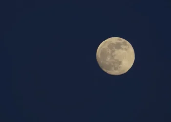 Luna piena quasi al completo nel cielo blu della sera, con i mari lunari scuri ben visibili sul disco