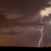 Maltempo: fulmini ramificati tra nubi scure sopra un'area di campagna piatta, con luce arancione all'orizzonte